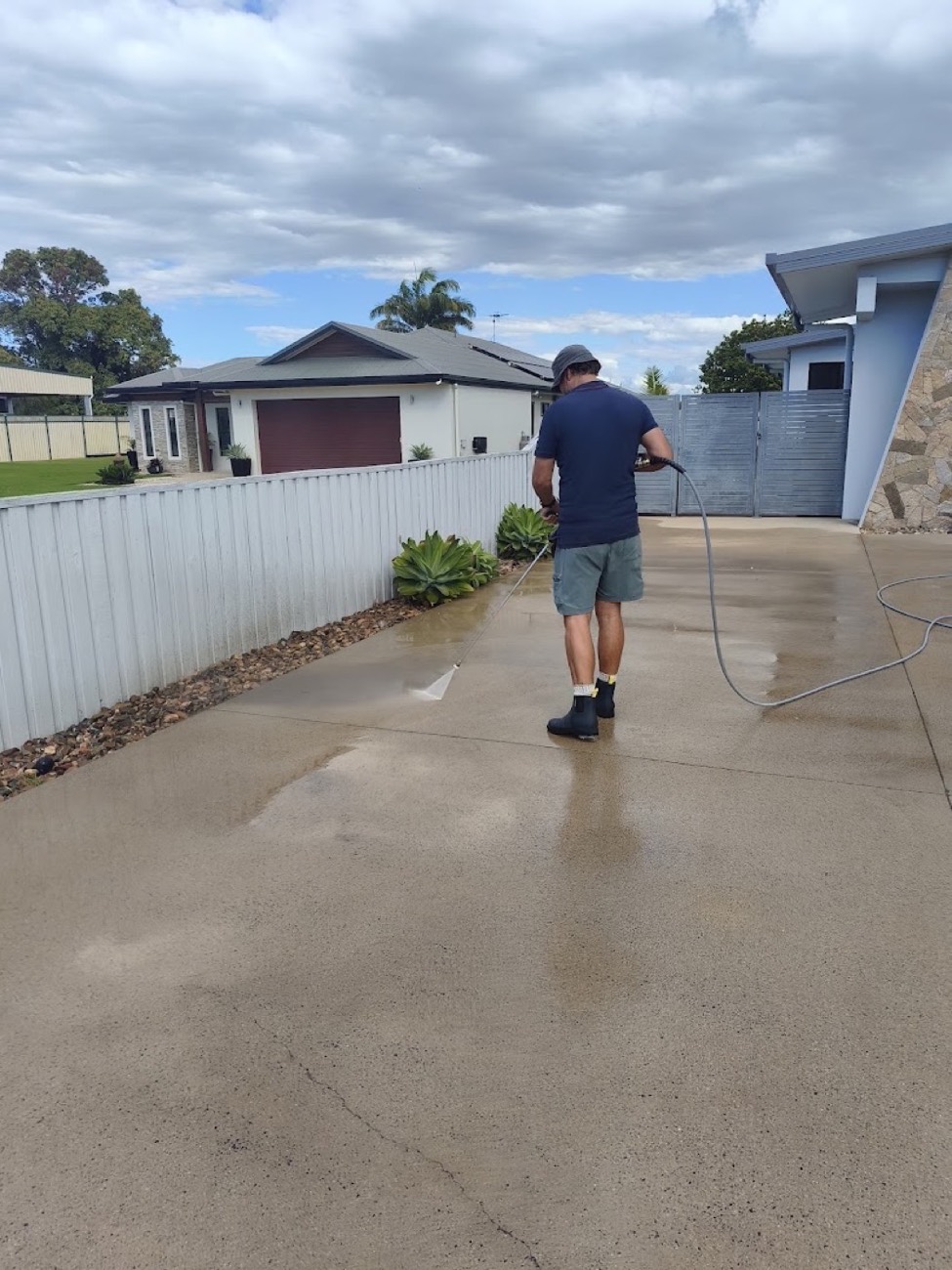 A man spraying water across a driveway beside a fence